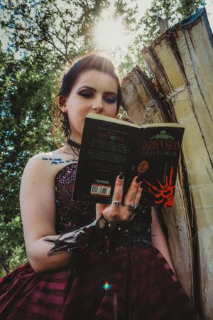 A person in a dark formal dress reads a book while posing against a stone wall outdoors.