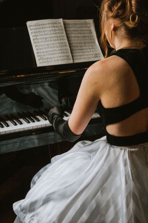 A pianist in a black top and white skirt reads sheet music while seated at a piano.