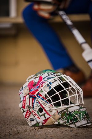A decorated hockey goalie mask sits on the ground with blue-suited legs in the background.