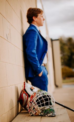Portrait of an athlete in a blue suit leaning against a wall with a hockey helmet on the ground.