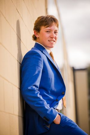 Portrait of a person in a bright blue suit and gold tie leaning against a beige wall.