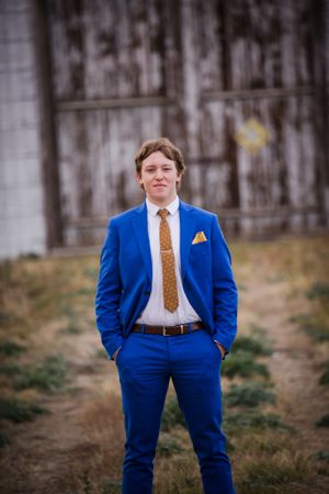 Formal portrait showing a person in a royal blue suit with gold tie against a rustic backdrop.