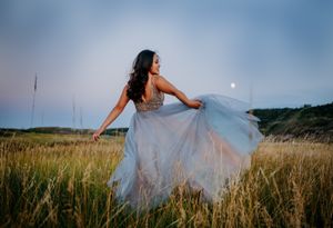 Woman in flowing gray dress twirling in golden field at sunset with moon visible.