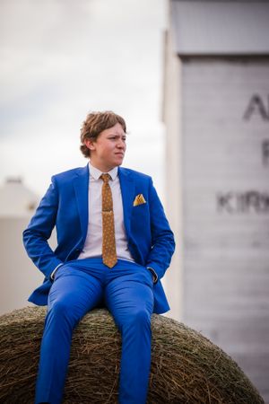 Person in bright blue suit sitting casually on hay bale against cloudy sky.