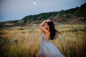 Woman in silver beaded gown dancing in tall grass during sunset.
