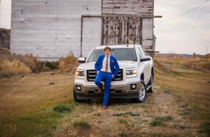 Person in bright blue suit poses casually against a white GMC truck near rustic barn.