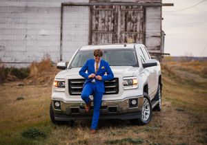 Person in bright blue suit leans against white GMC truck with weathered barn in background.