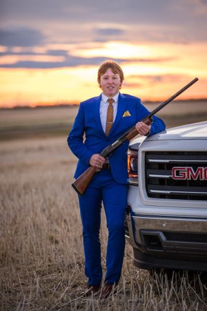 Figure in royal blue suit poses with hunting rifle against sunset sky and GMC truck in rural field.