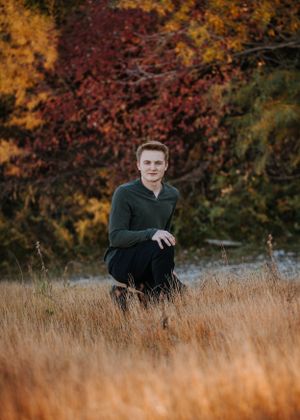 Portrait of person kneeling in tall grass with vibrant fall colors in background.