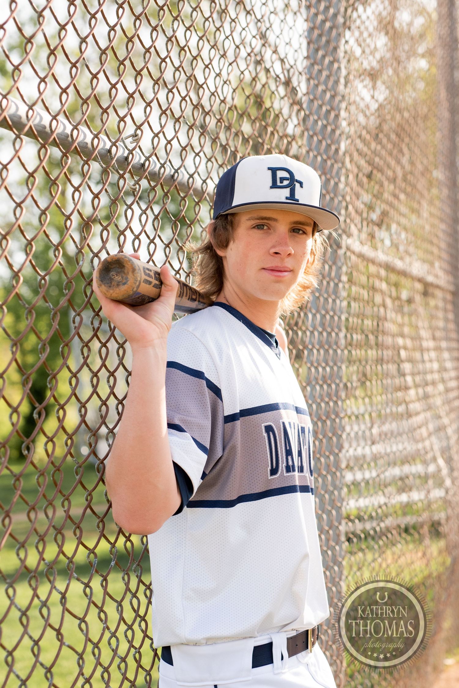 Baseball player in white Dallas uniform poses with bat against chain link fence.
