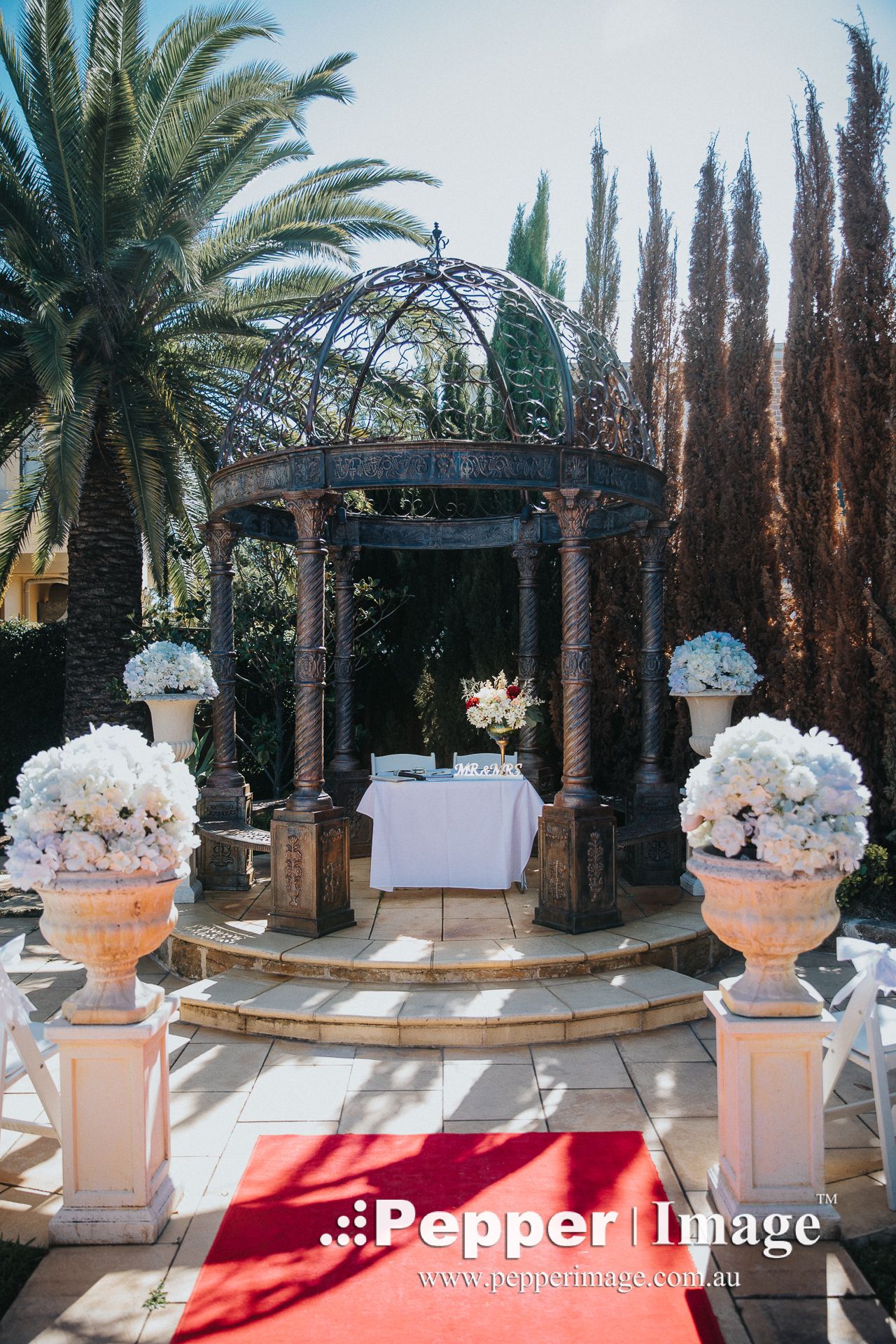 Elegant outdoor wedding ceremony setup with a wrought iron gazebo, red carpet, and white floral urns.