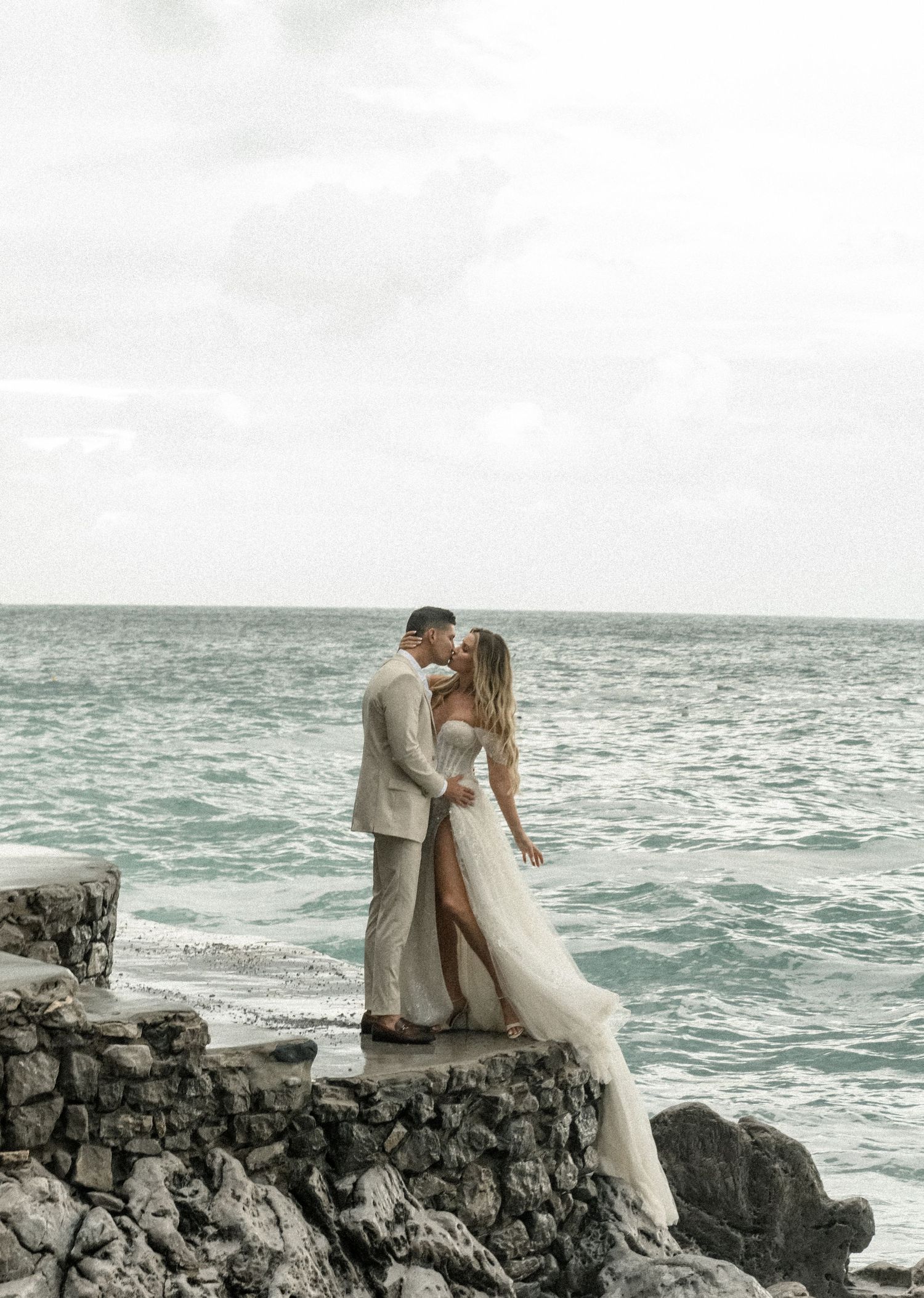 A romantic wedding couple embraces on rocky cliffs overlooking turquoise waters at sunset.
