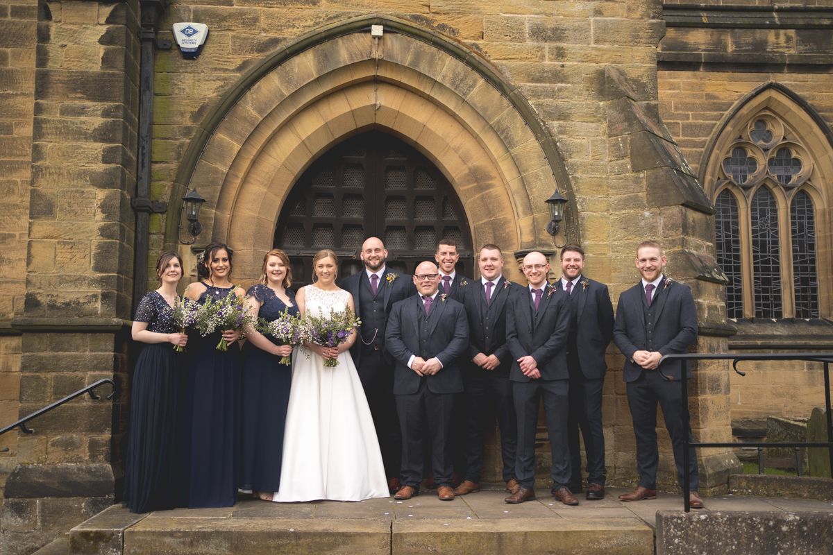 Wedding party poses for formal group photo outside historic stone church entrance.