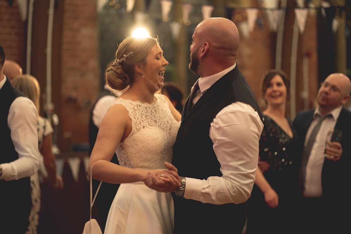 A couple shares a joyful dance moment at their wedding reception in a rustic brick venue with string lights.