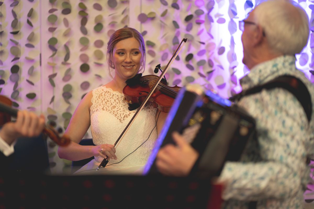 Musicians perform at a wedding reception with violin and accordion under purple lighting.