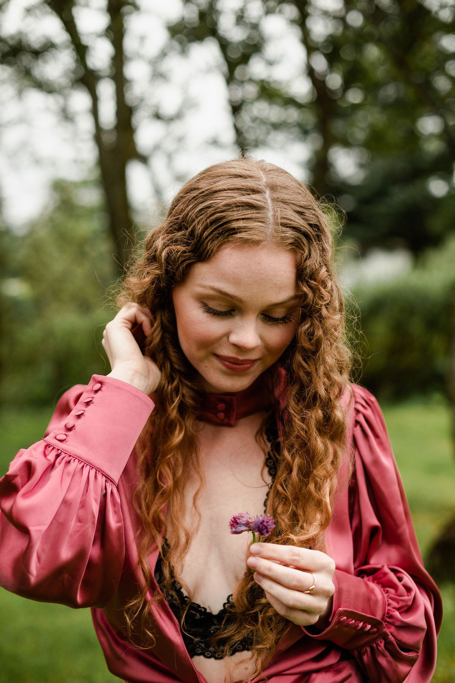 Woman in pink dress admires wildflower in lush green garden setting.