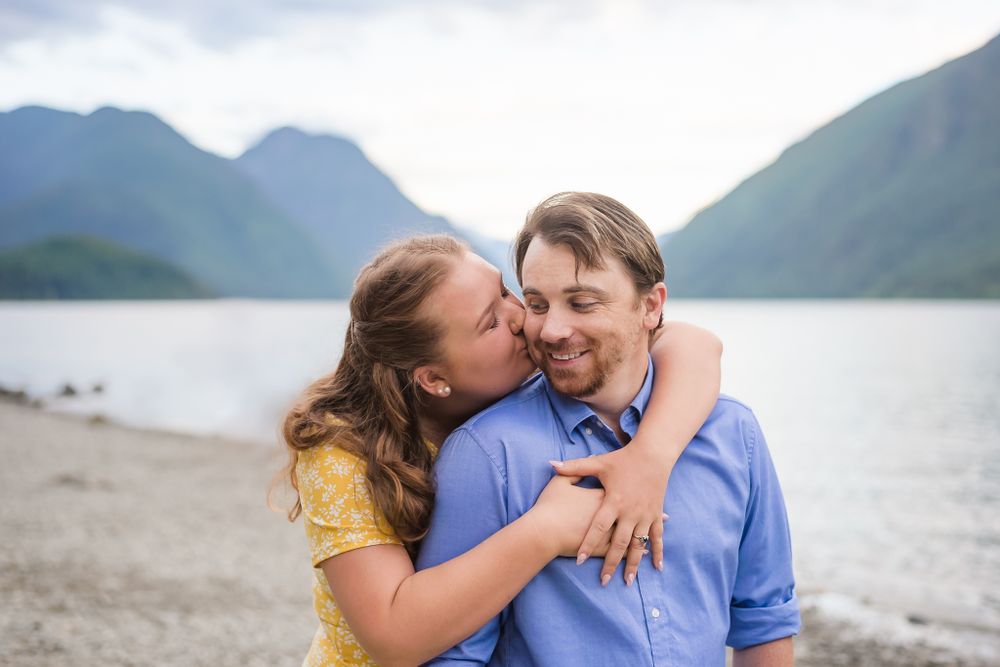 A woman wraps her arms around her fiance from behind and kisses him on the cheek.