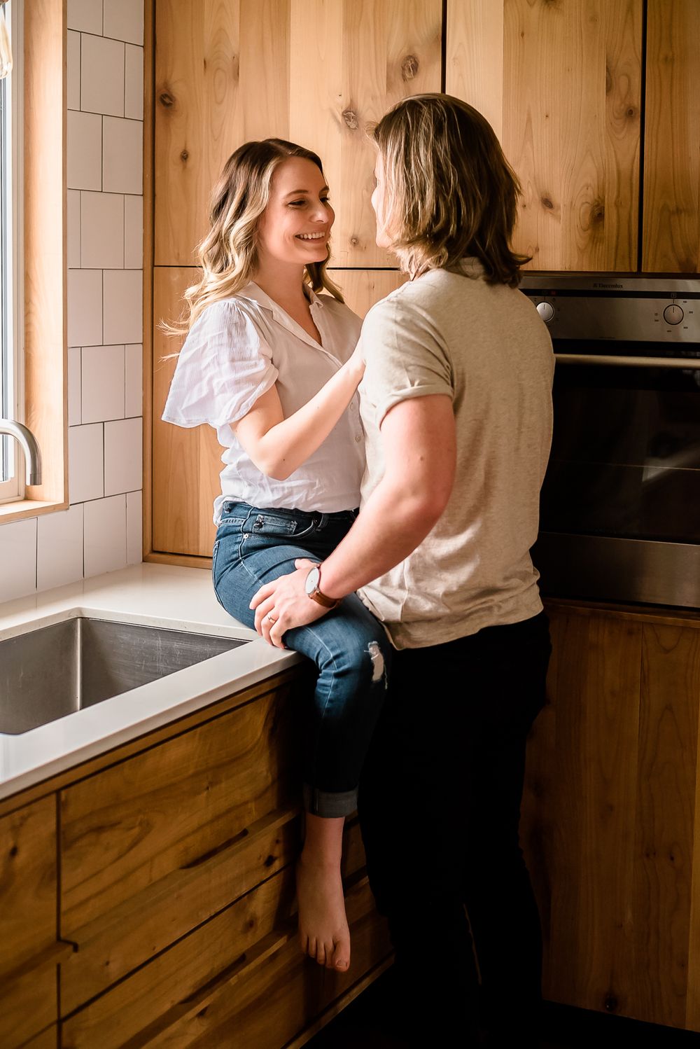 A man and woman look in to each other's eyes while the woman sits on the kitchen counter. Natural light floods the kitchen around them.