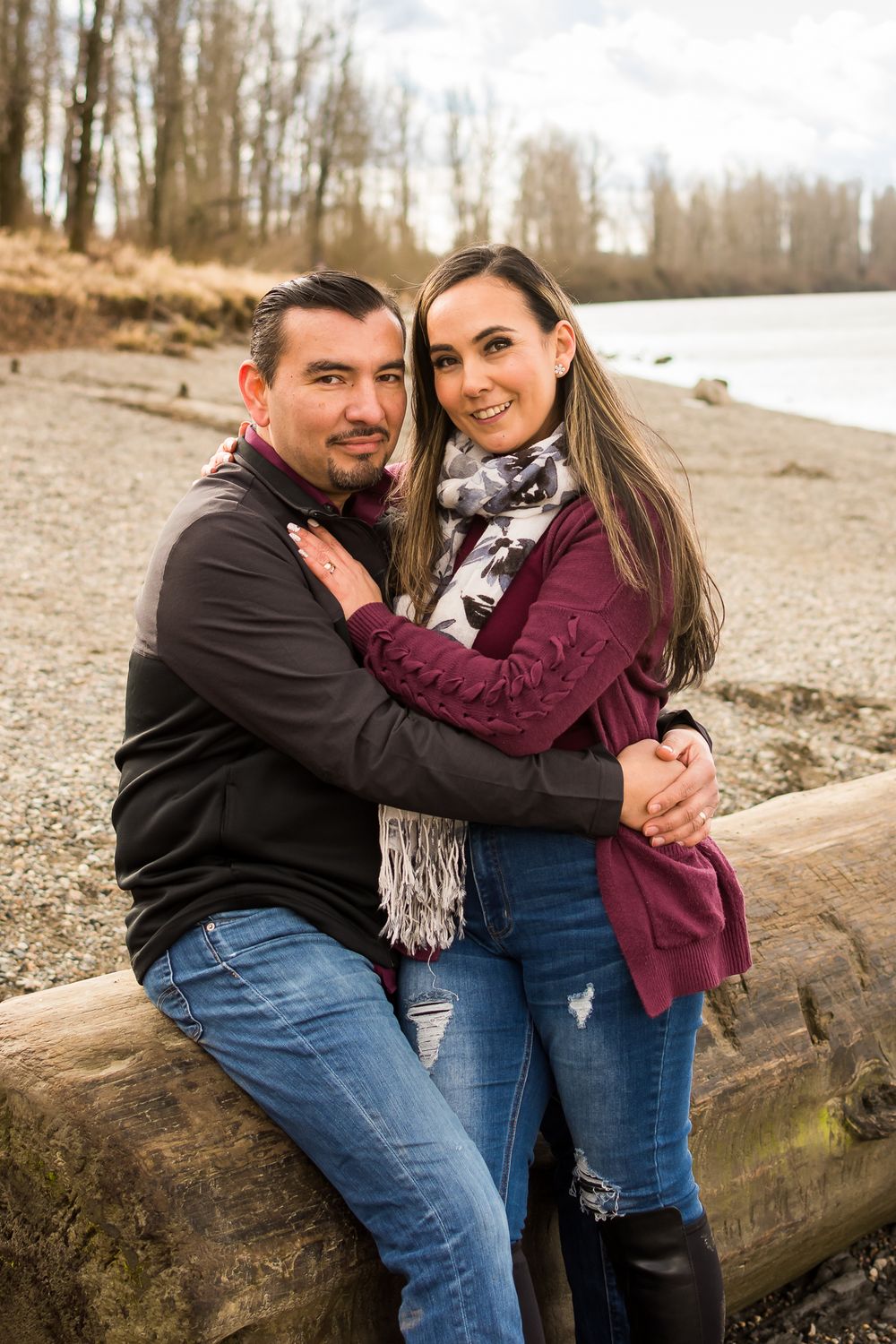 On a winter afternoon, a couple stand with their arms around one another with the river and trees behind them.