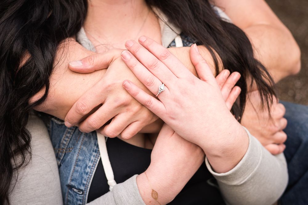 A close up shot of a woman with her husband's arms around her. She is wearing a bracelet and has an engagement ring on her finger.