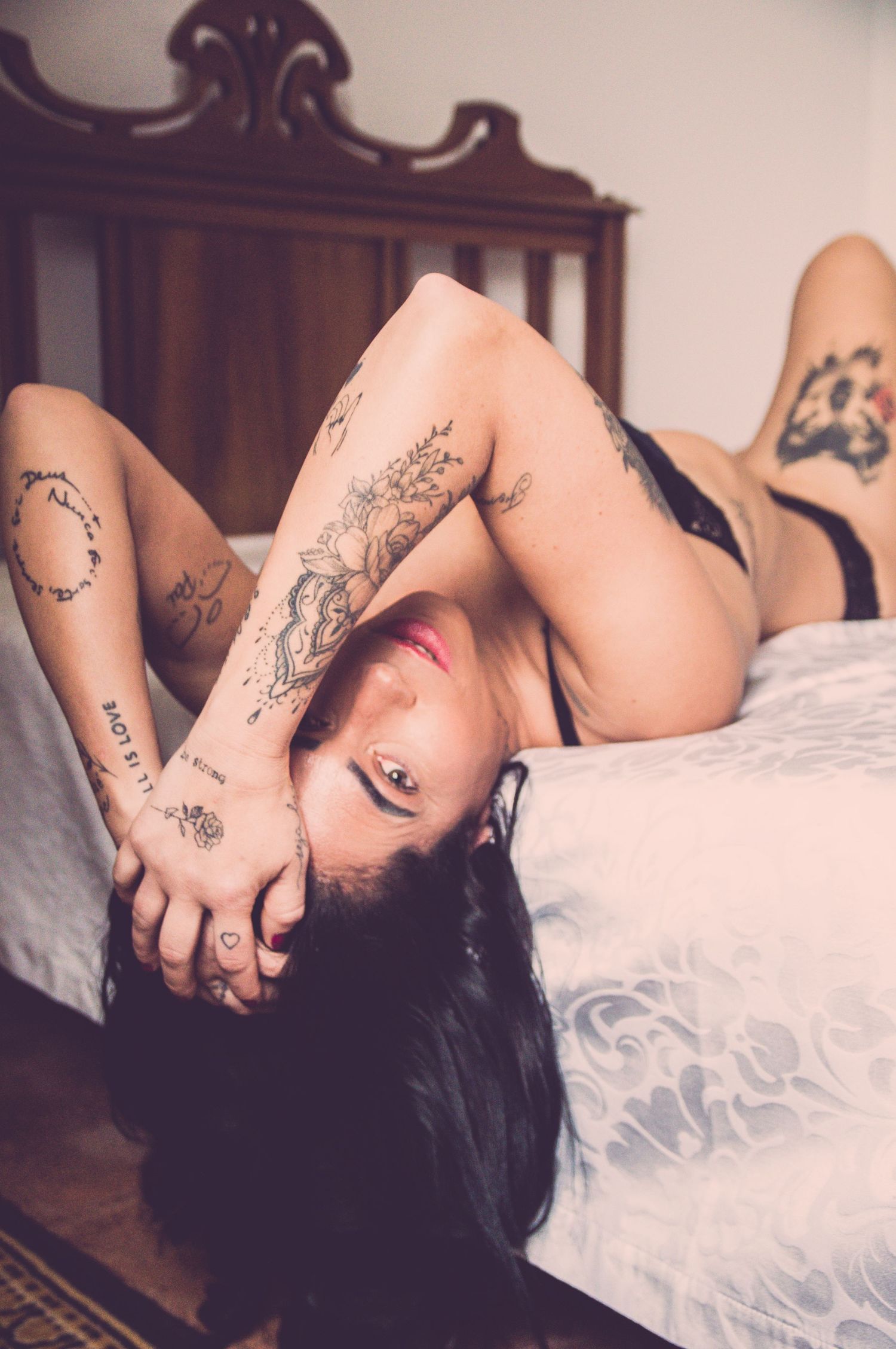 Person with tattoos lying on white bedding in dimly lit room.