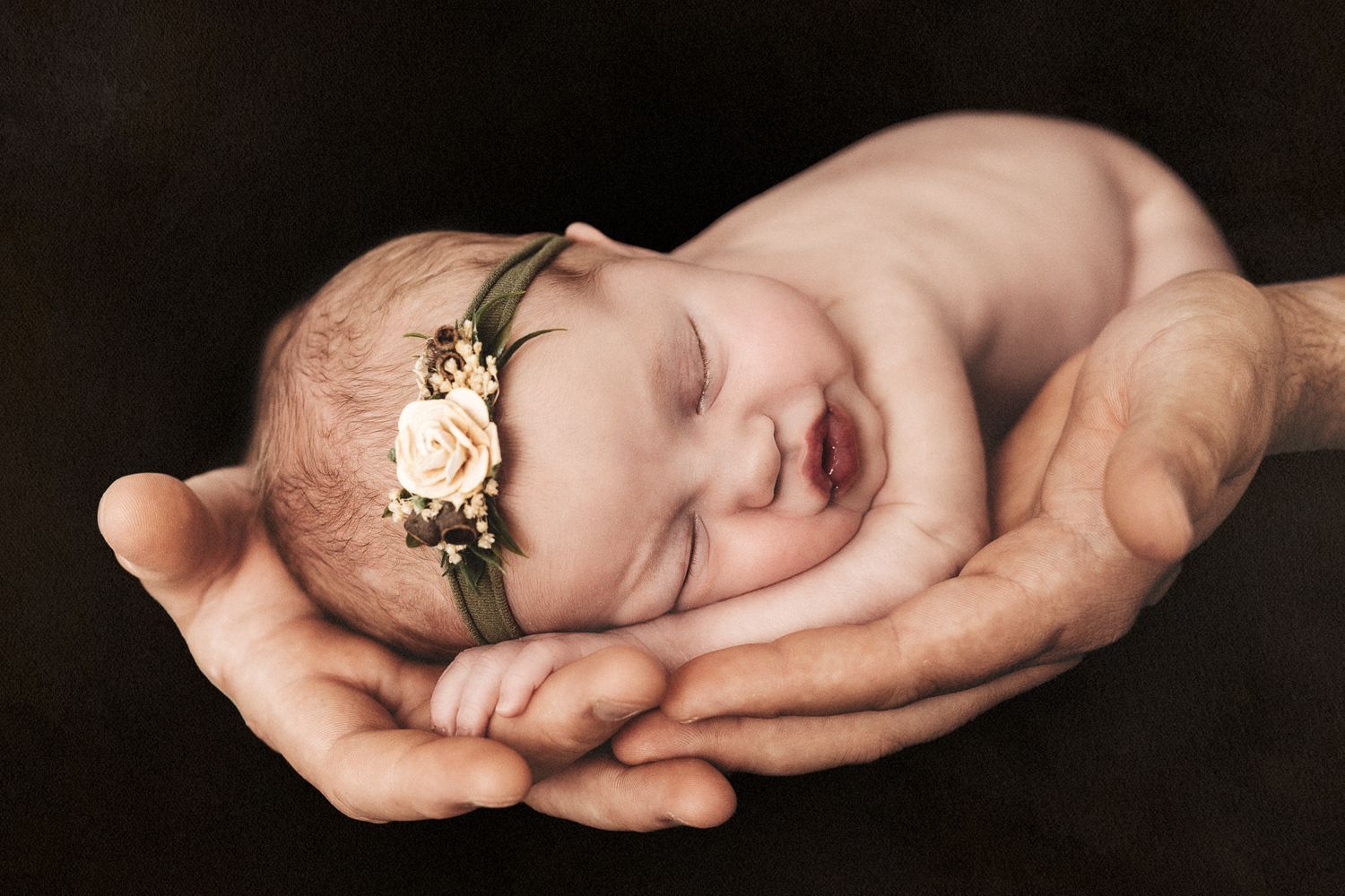 Sleeping newborn with floral headband cradled gently in hands against dark background.