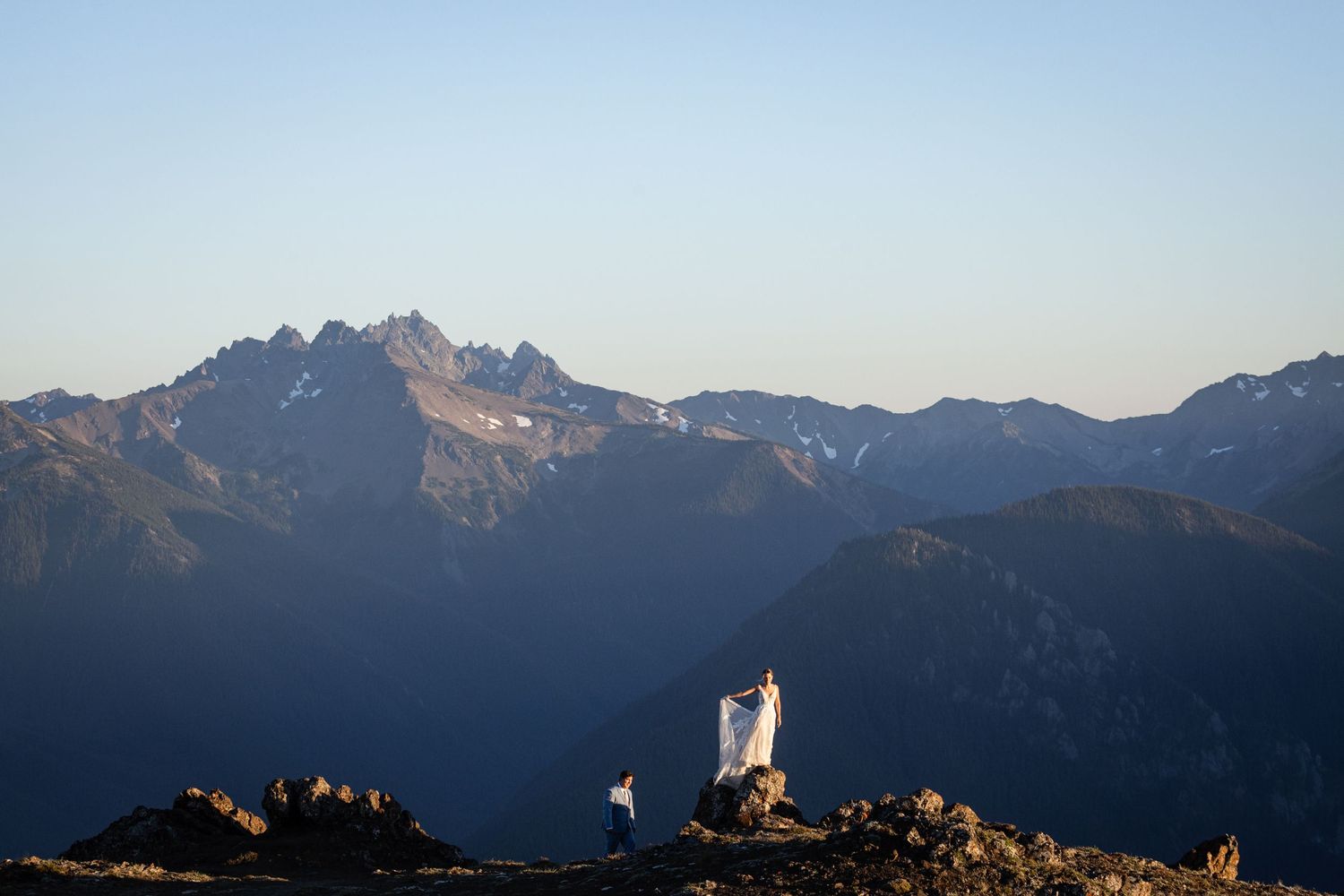 Lone bride in billowing white wedding gown stands on mountain summit overlooking dramatic valley below.
