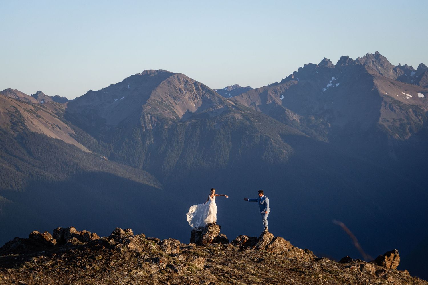 Bride in flowing white gown dances with groom on rocky mountain summit with vast peaks backdrop.