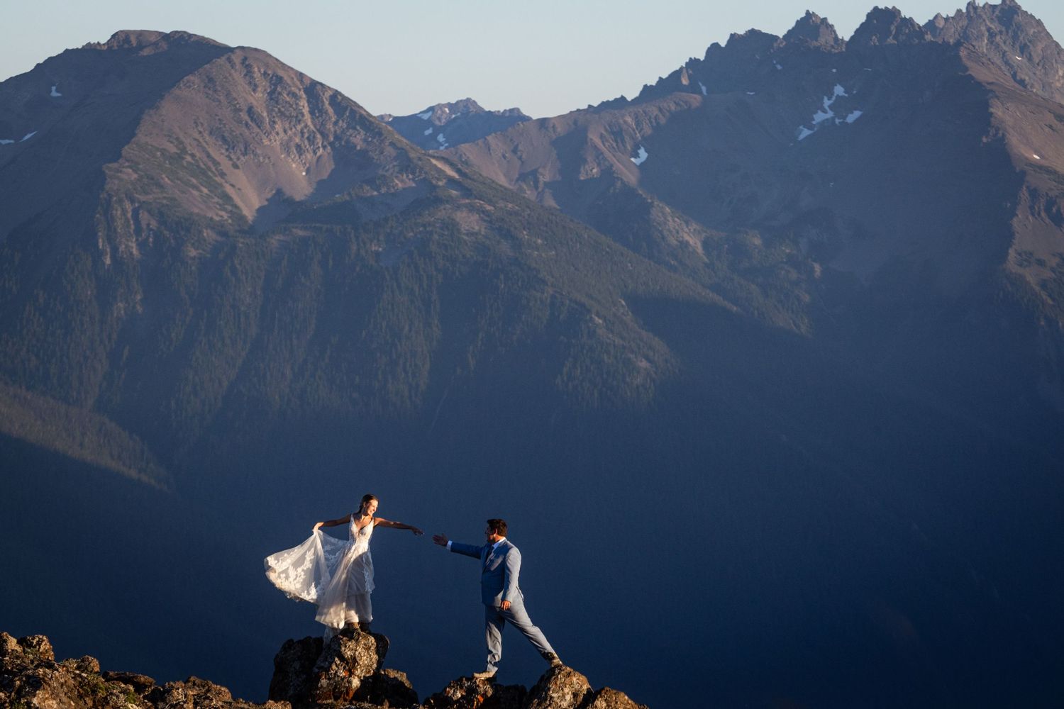 Bride and groom hold hands on rocky mountain peak with stunning layered mountain range behind them.