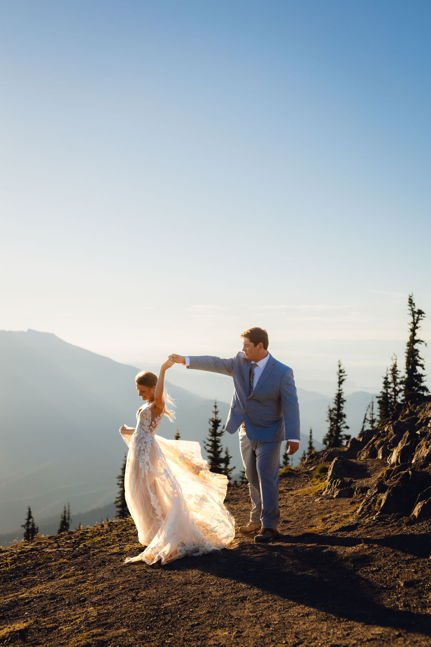 Groom twirls bride in a flowing lace gown on a mountain summit with breathtaking valley views.