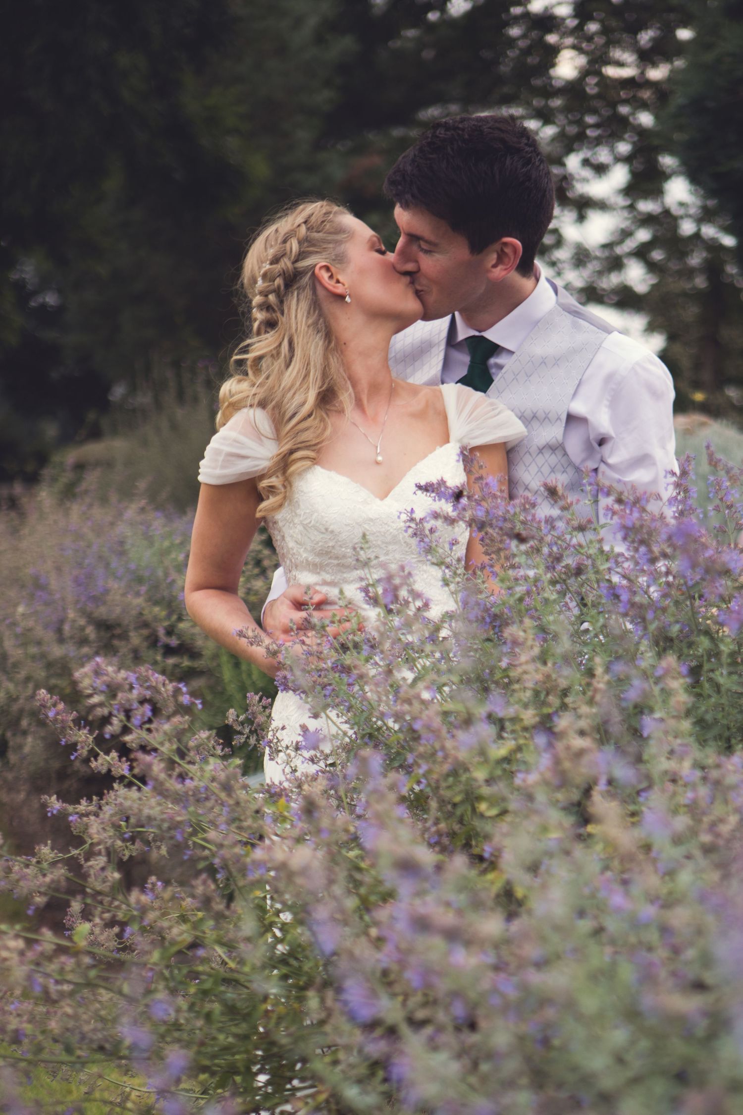 Bride and groom kissing with purple flowers in foreground on wedding day during couples photography session