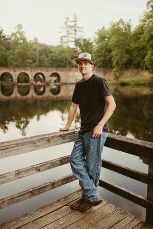 Person standing on wooden deck overlooking scenic stone bridge and pond surrounded by trees.