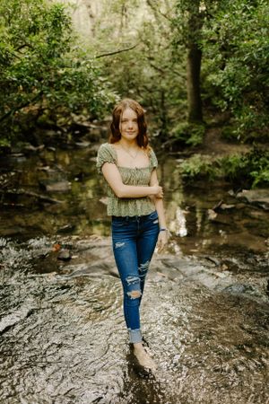 Person in green top and ripped jeans stands confidently on rocks in creek.