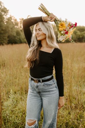 Woman in black top and ripped jeans holds colorful wildflower bouquet in golden field at sunset.