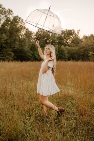 Woman in white dress twirls with clear umbrella in golden field.