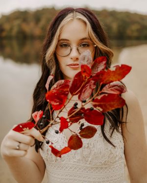 Artistic portrait with red autumn leaves and white lace dress against a lake backdrop.