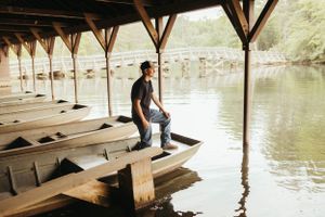 Person sits contemplatively on wooden dock overlooking tranquil lake with wooden support beams overhead.