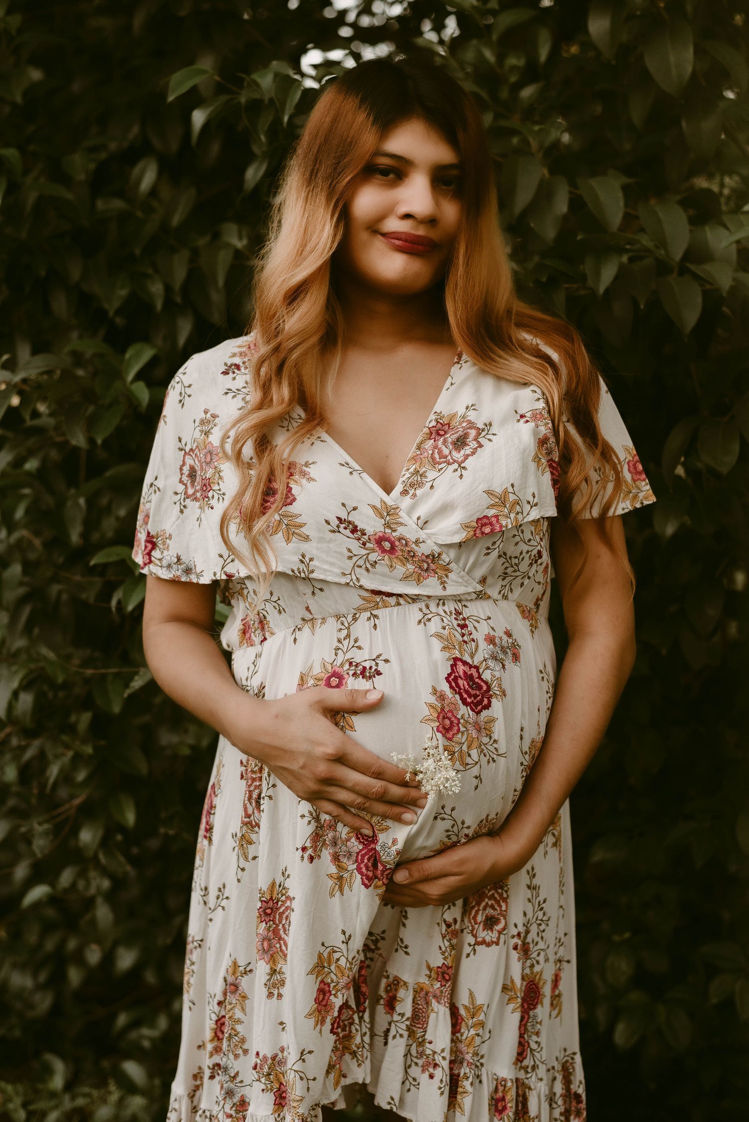 An expectant parent poses in a floral wrap dress outdoors with greenery in the background.