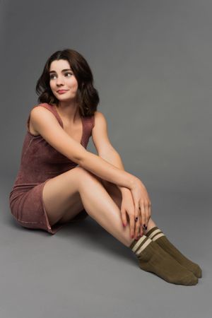 High School senior portrait of a girl in a brown dress sits on studio floor wearing olive green knee socks against gray background.