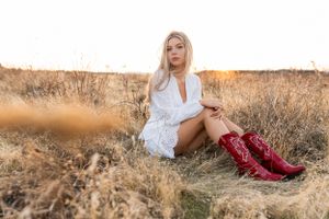 High school senior photos of a girl in a white dress and red cowboy boots sits in a golden field during sunset.