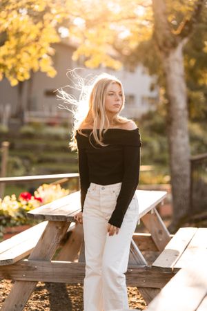 Golden autumn sunlight illuminates high school senior girl in black off-shoulder top and white pants posing on wooden picnic table.