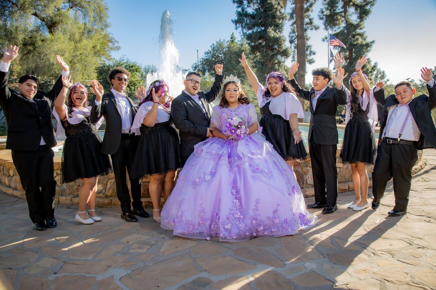 Quinceañera girl in lavender ball gown celebrates with her court outdoors near a fountain.