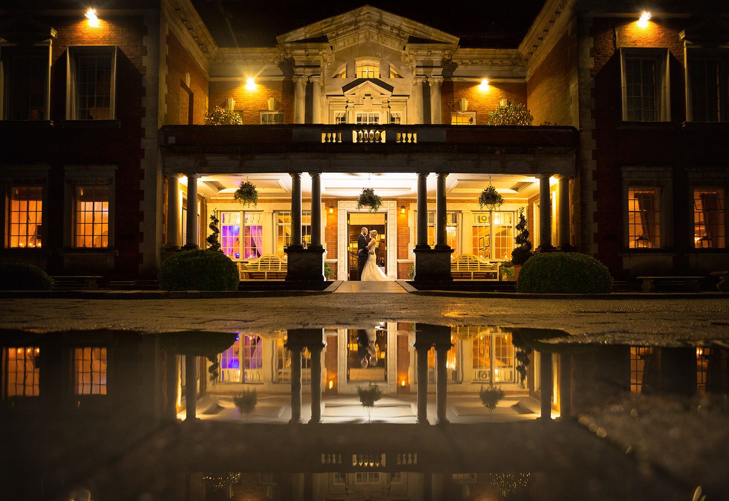 Bride and groom embrace and are reflected in a huge rain puddle before the building of Eaves Hall wedding venue