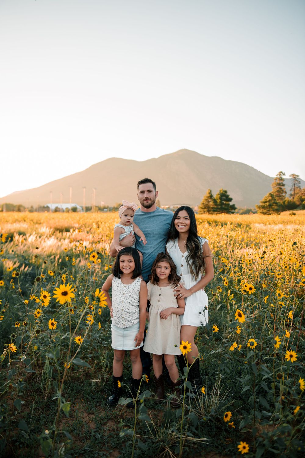 Family portrait in a vibrant field of sunflowers with mountains and evergreen trees at golden hour.