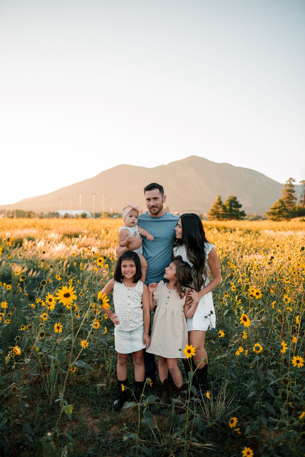 Family poses together in a sunflower meadow with mountain backdrop during sunset.