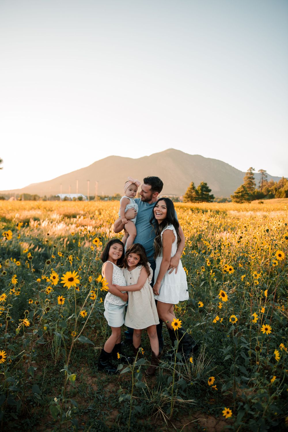 Family group photo in field of yellow sunflowers with mountain landscape behind them.