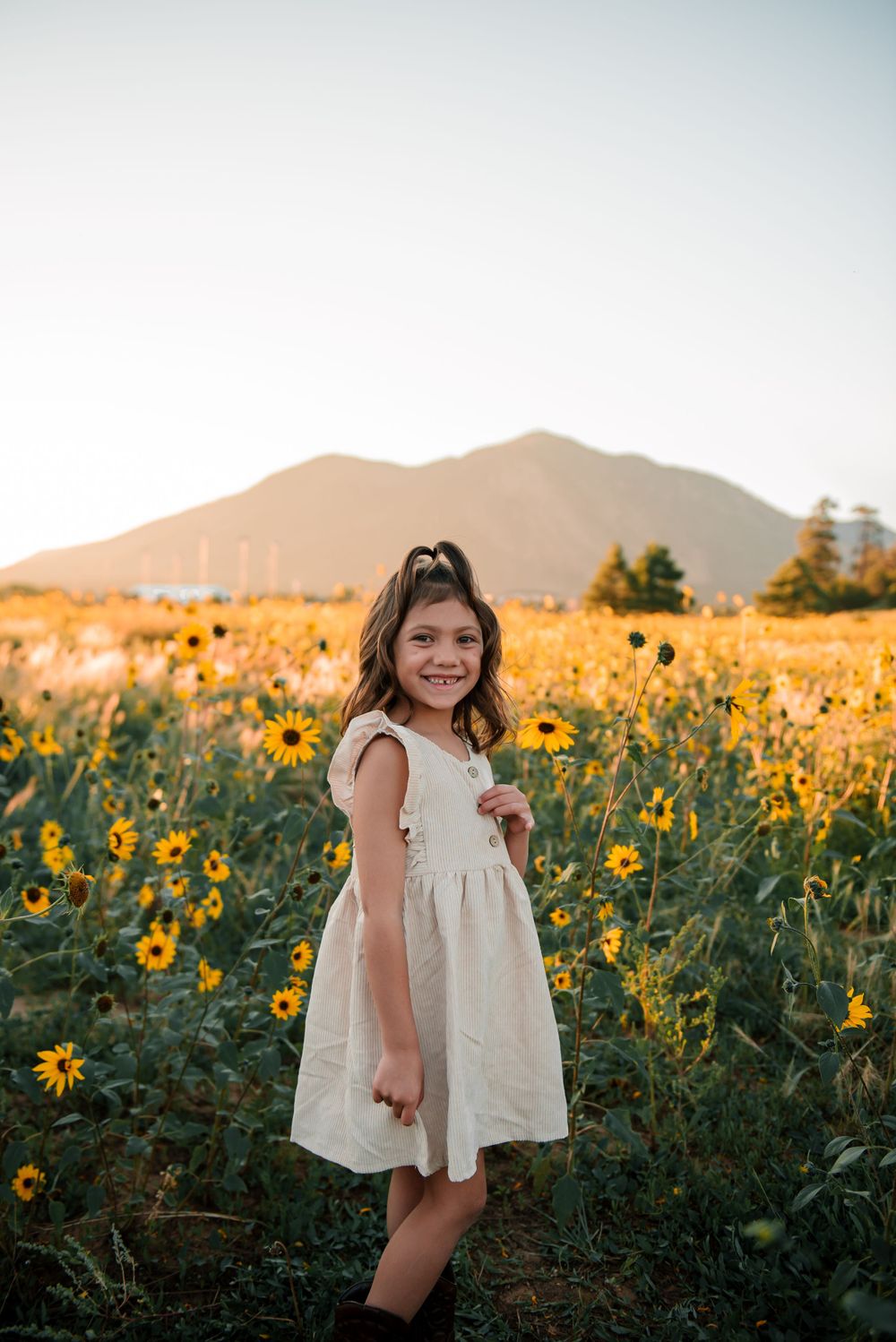 Young girl in white dress smiling in sunflower field with mountain backdrop at sunset.