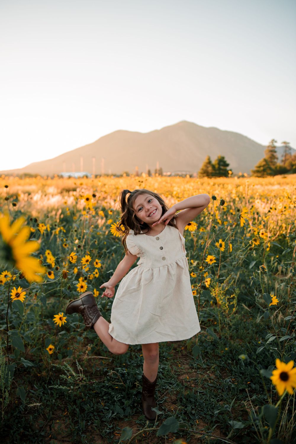 Young girl twirling in white dress among sunflowers with mountains in background at sunset.