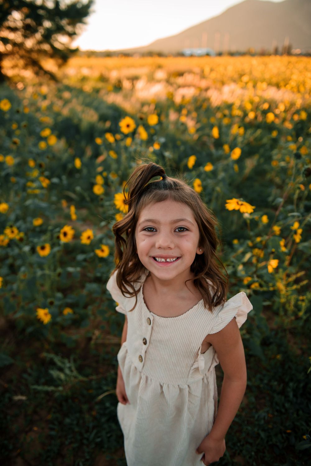 A child in a white dress smiles joyfully in a sunflower field during golden hour.