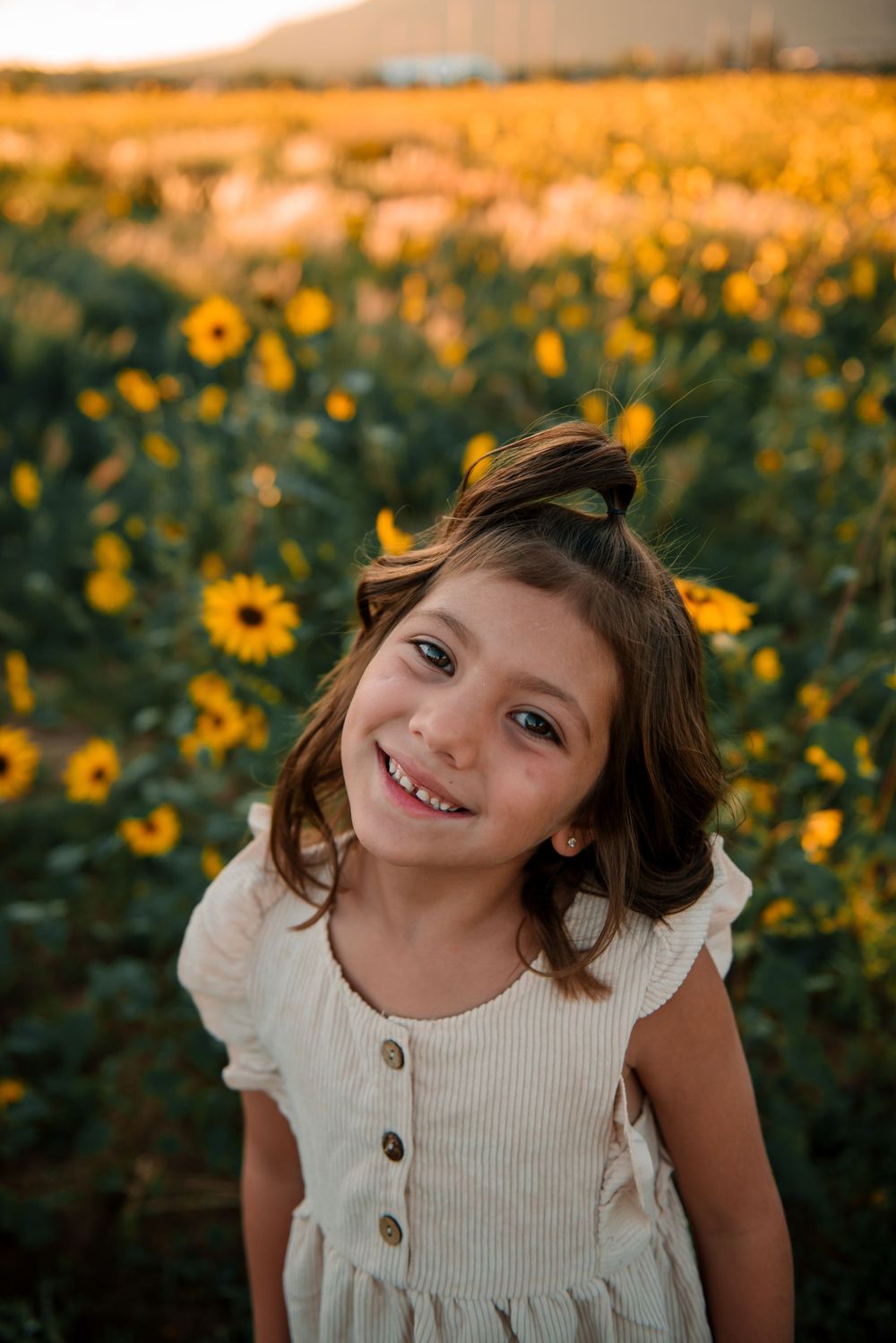 A child poses playfully in a white dress among blooming sunflowers at sunset.